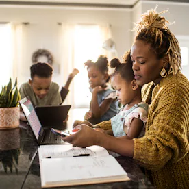 A mother helps her young daughter with homework at a kitchen island while two other children use lap...