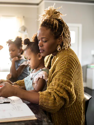 A mother helps her young daughter with homework at a kitchen island while two other children use lap...
