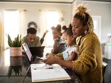 A mother helps her young daughter with homework at a kitchen island while two other children use lap...