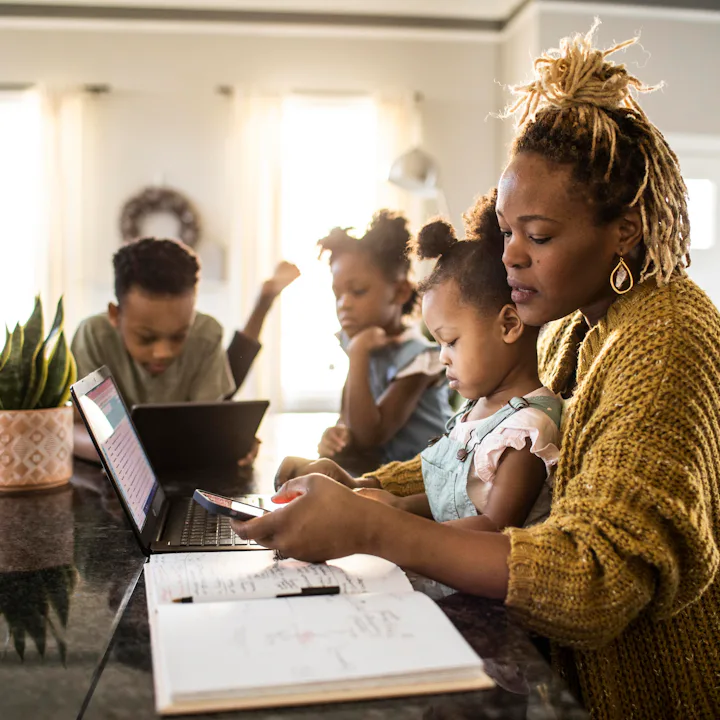 A mother helps her young daughter with homework at a kitchen island while two other children use lap...
