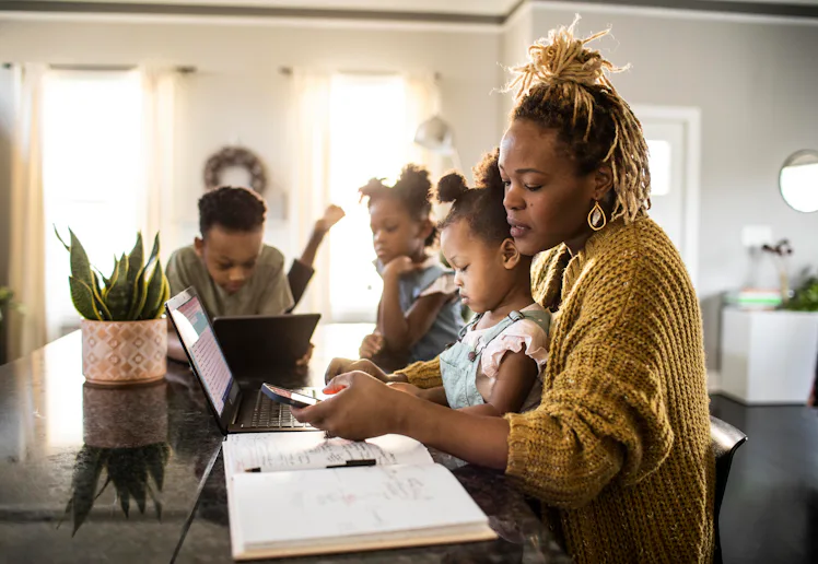 A mother helps her young daughter with homework at a kitchen island while two other children use lap...
