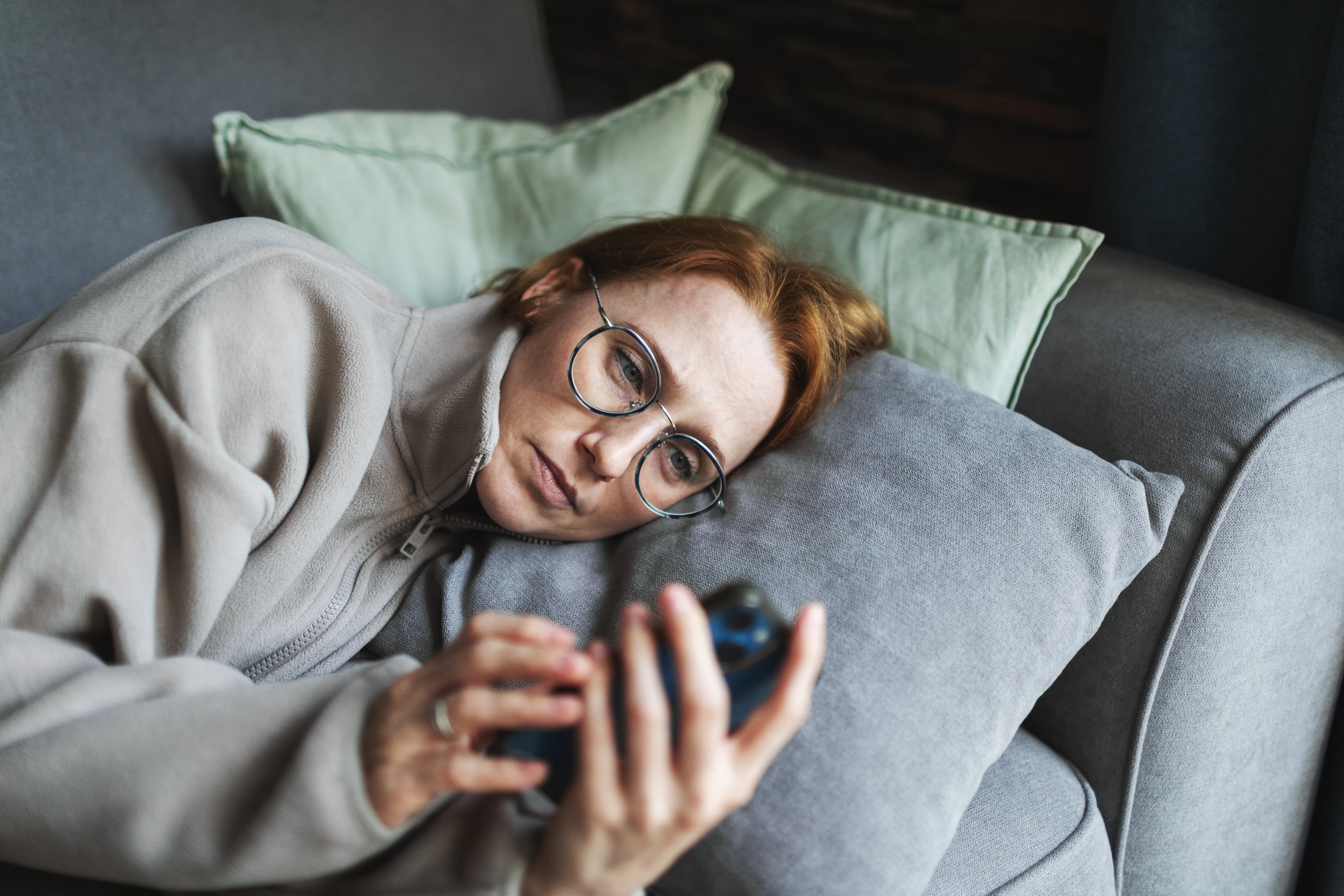 Person with glasses lying on a couch, propped on pillows, looking intently at a smartphone held in t...