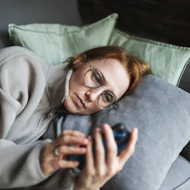 Person with glasses lying on a couch, propped on pillows, looking intently at a smartphone held in t...
