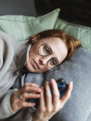 Person with glasses lying on a couch, propped on pillows, looking intently at a smartphone held in t...