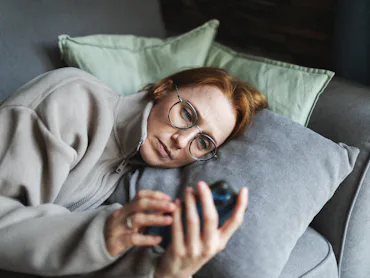 Person with glasses lying on a couch, propped on pillows, looking intently at a smartphone held in t...