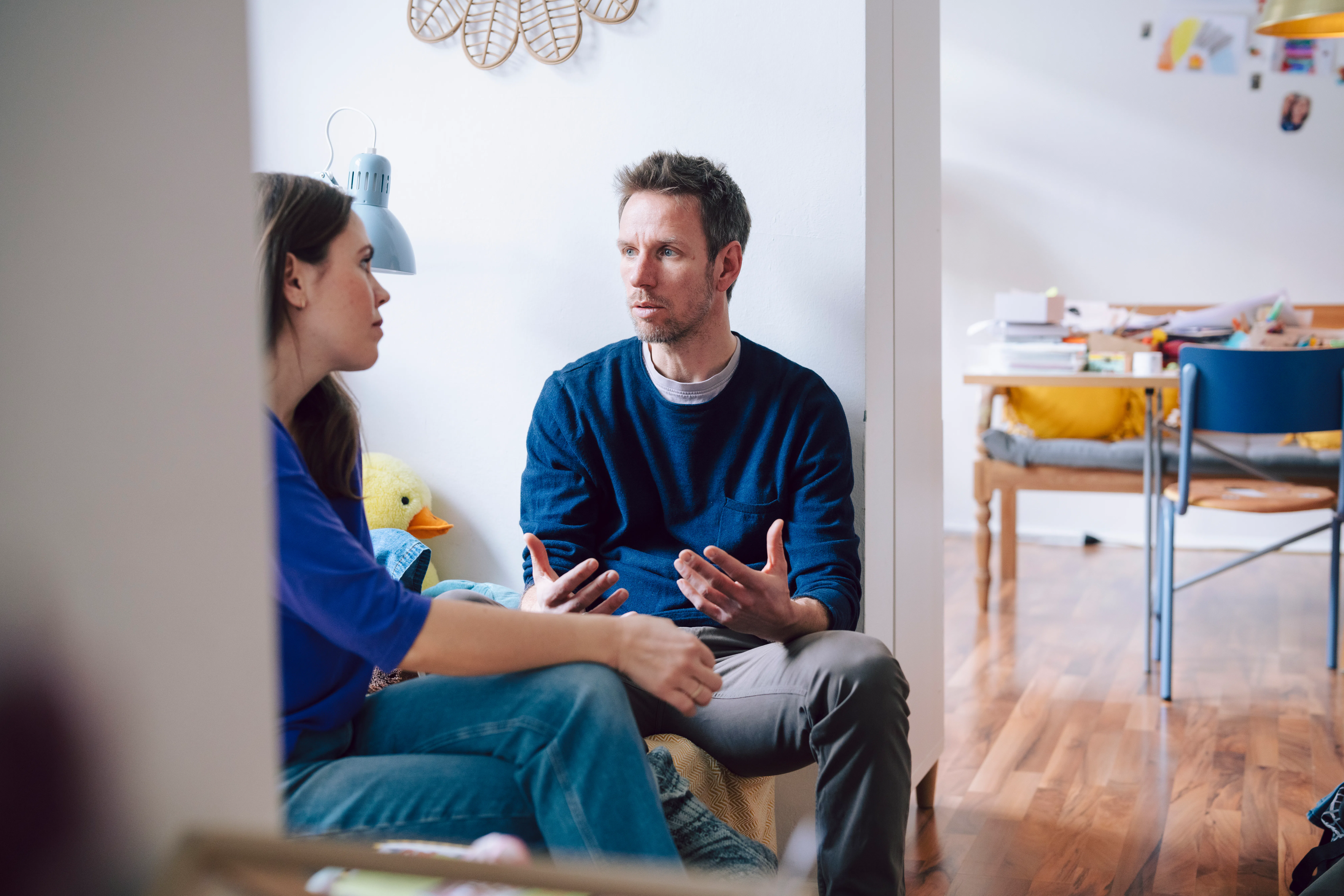 A man and woman sit facing each other in a living area, engaged in a serious conversation; a child's...