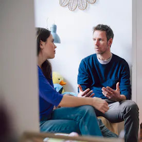 A man and woman sit facing each other in a living area, engaged in a serious conversation; a child's...