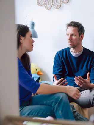 A man and woman sit facing each other in a living area, engaged in a serious conversation; a child's...