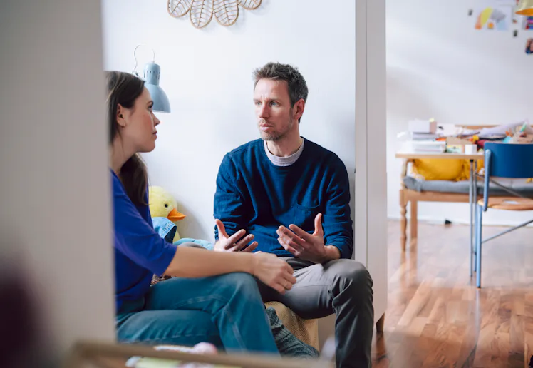 A man and woman sit facing each other in a living area, engaged in a serious conversation; a child's...