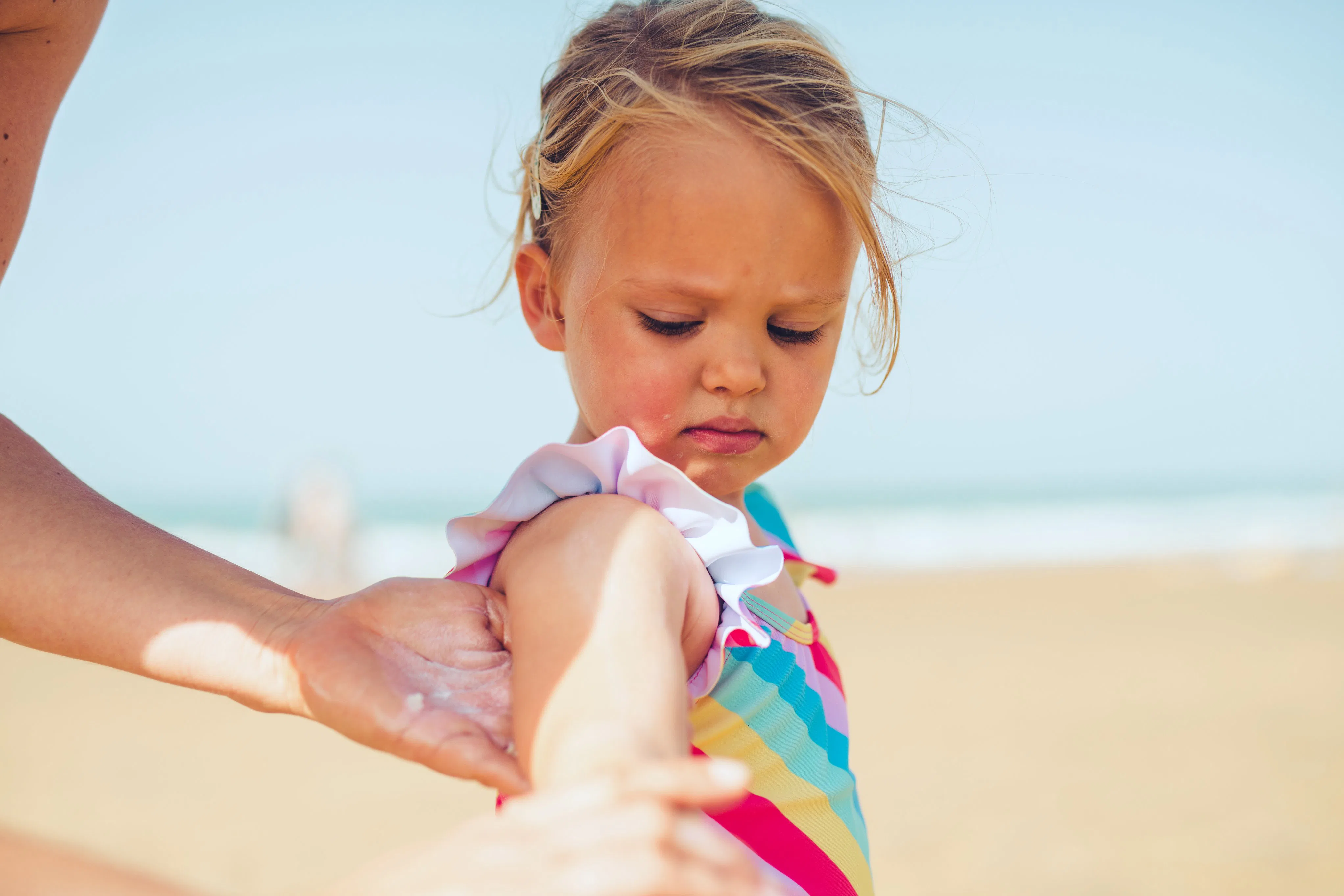 Young child in a colorful striped swimsuit standing on a sandy beach while an adult hand applies sun...