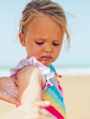 Young child in a colorful striped swimsuit standing on a sandy beach while an adult hand applies sun...