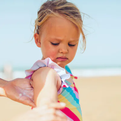 Young child in a colorful striped swimsuit standing on a sandy beach while an adult hand applies sunscreen to the child's upper arm.