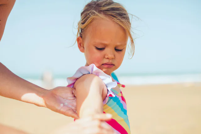 Young child in a colorful striped swimsuit standing on a sandy beach while an adult hand applies sun...