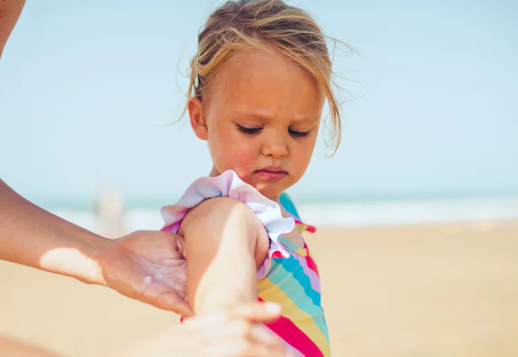 Young child in a colorful striped swimsuit standing on a sandy beach while an adult hand applies sun...
