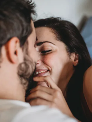 Close-up of a smiling couple cuddling on a couch, woman leaning in toward a bearded man with hands g...