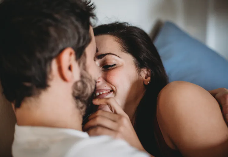 Close-up of a smiling couple cuddling on a couch, woman leaning in toward a bearded man with hands g...