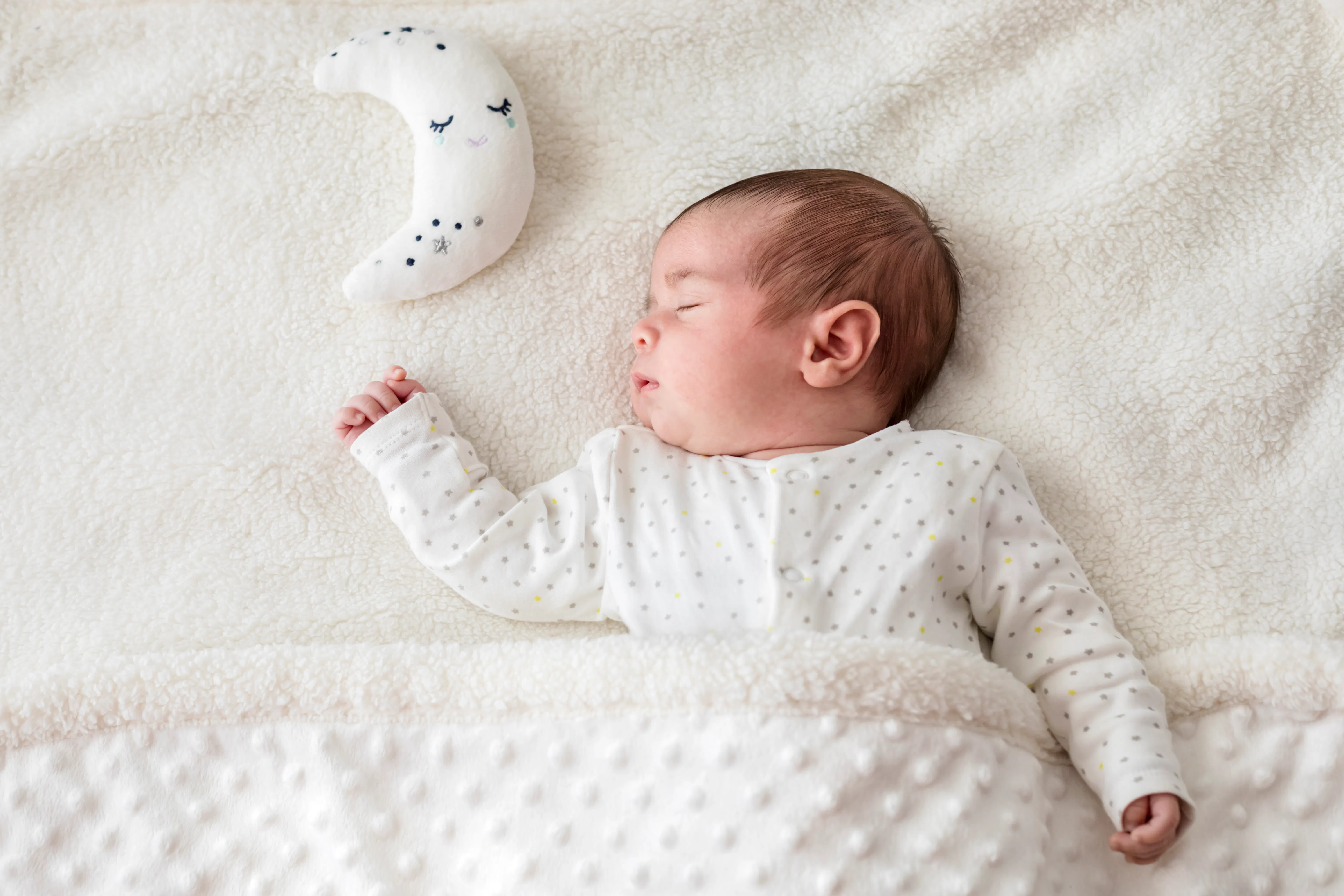 Sleeping newborn baby swaddled in a dotted white onesie under a textured blanket, lying on a soft cr...