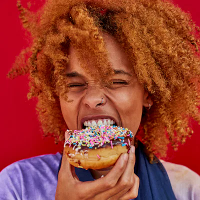Young person with curly hair biting a pink-frosted sprinkled donut, eyes closed and smiling, holding it against a bright red background.