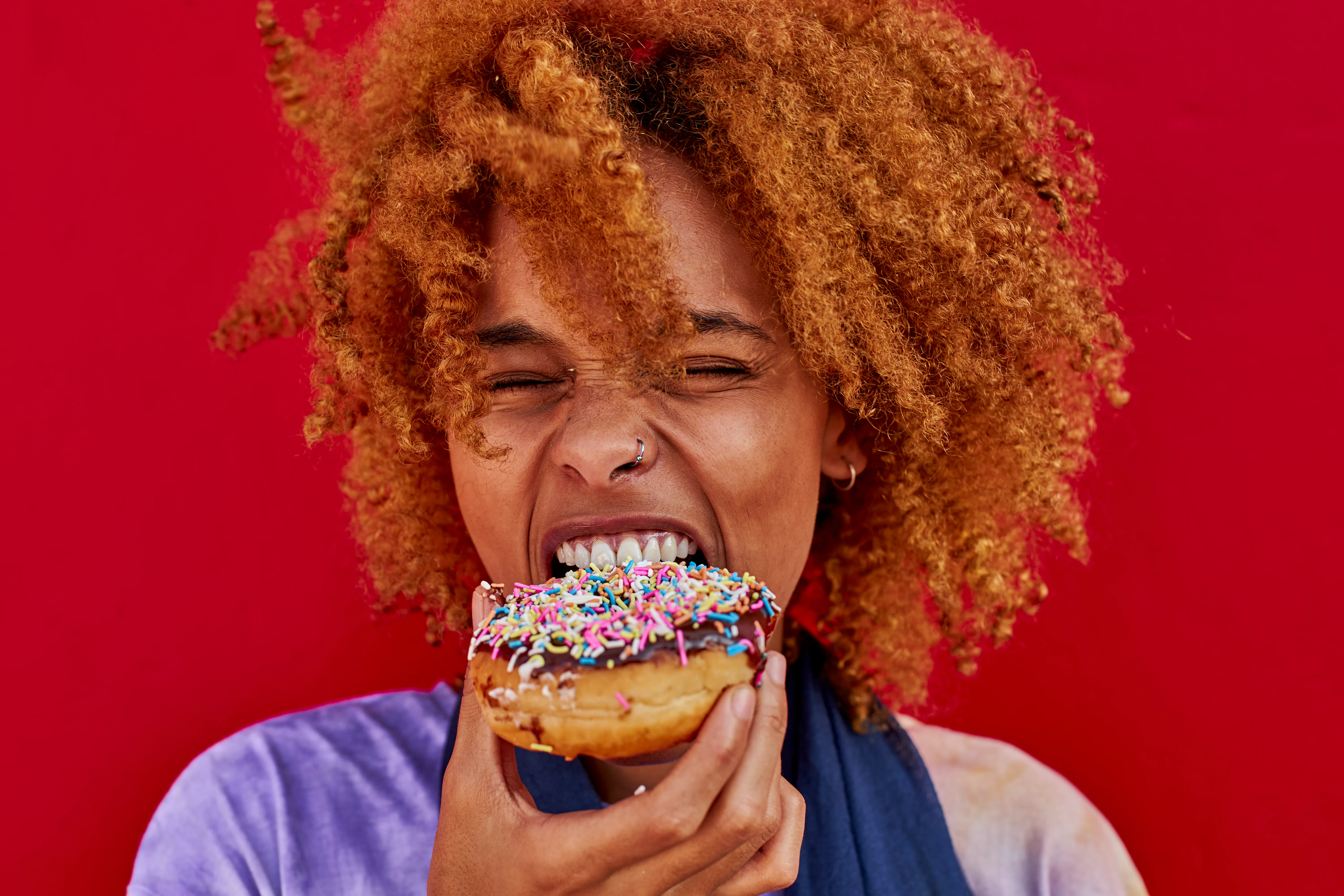 Person with curly reddish hair and nose ring biting a sprinkled-glazed donut, holding it near their ...