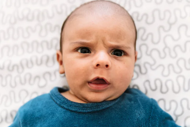 Close-up of a confused-looking infant lying on a patterned sheet, wearing a blue shirt, with furrowe...