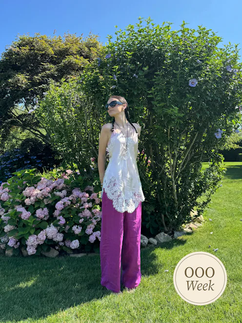 Woman outside in front of foliage wearing purple linen pants and white blouse