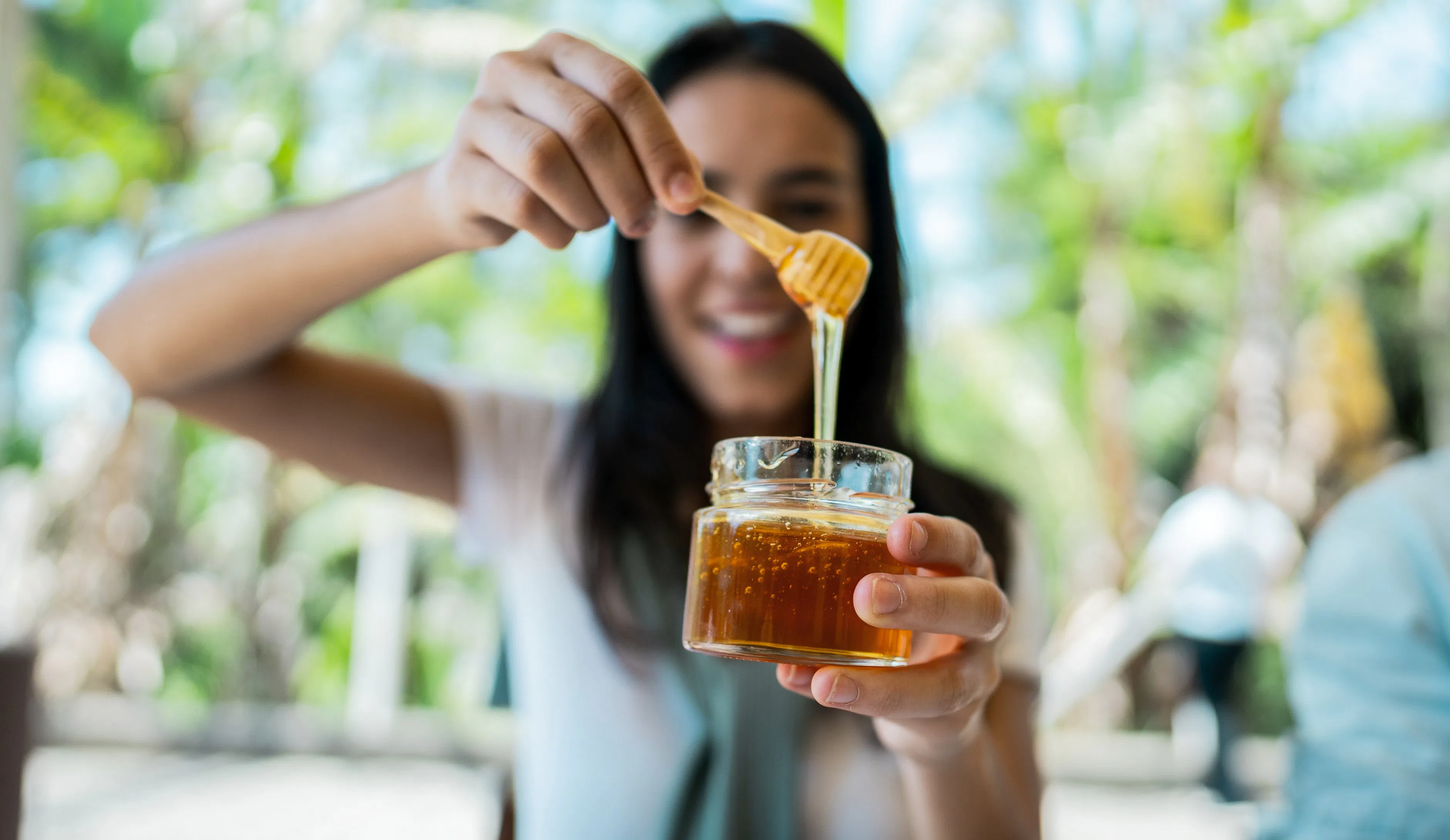 Smiling woman holding a small glass jar of honey with a wooden dipper, honey dripping from dipper ag...