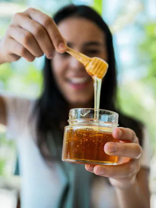 Smiling woman holding a small glass jar of honey with a wooden dipper, honey dripping from dipper ag...