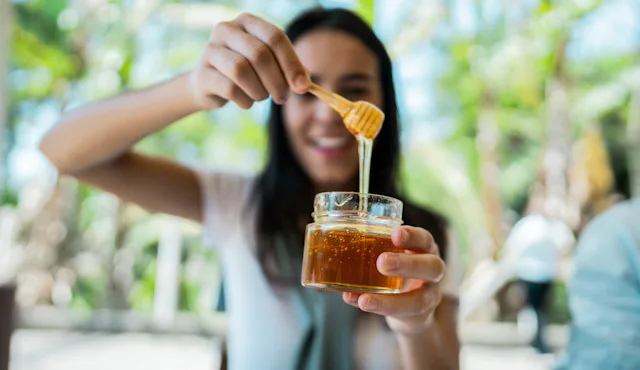 Smiling woman holding a small glass jar of honey with a wooden dipper, honey dripping from dipper ag...