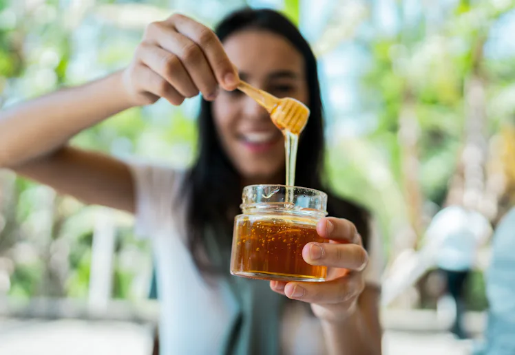 Smiling woman holding a small glass jar of honey with a wooden dipper, honey dripping from dipper ag...