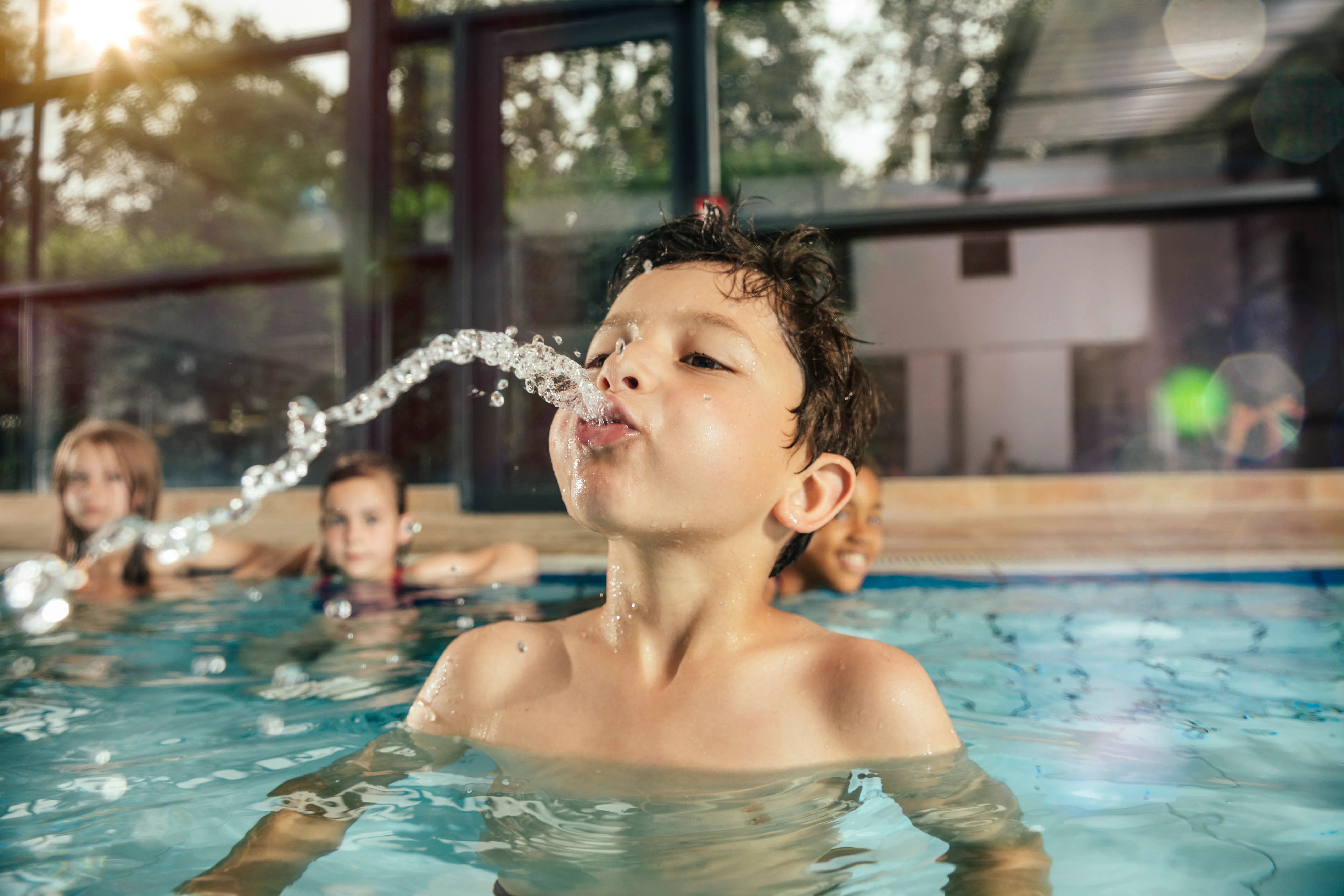 A young boy in an indoor pool leans back, spouting a stream of water from his mouth as three childre...