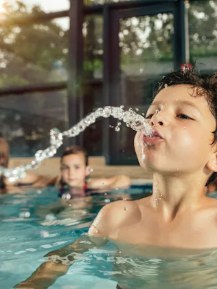A young boy in an indoor pool leans back, spouting a stream of water from his mouth as three childre...