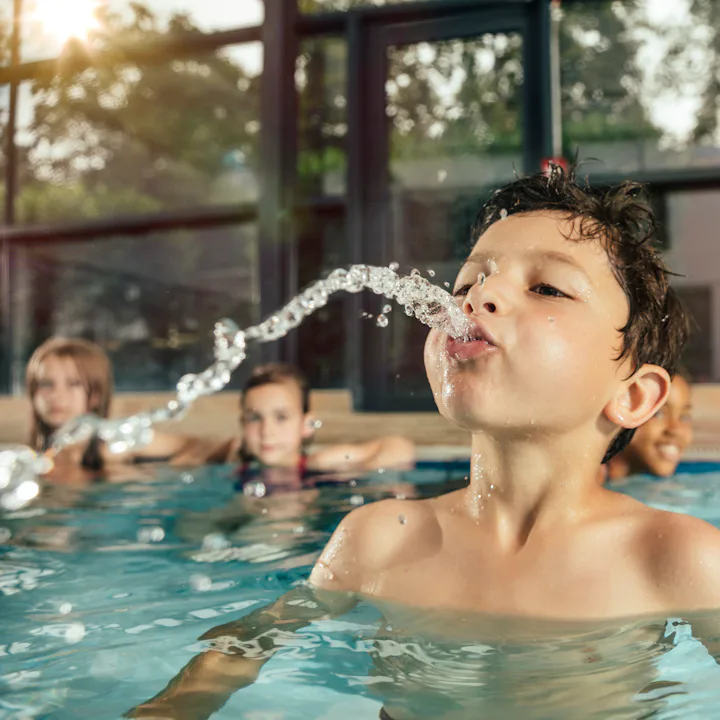A young boy in an indoor pool leans back, spouting a stream of water from his mouth as three childre...