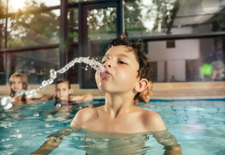 A young boy in an indoor pool leans back, spouting a stream of water from his mouth as three childre...