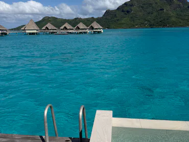 Turquoise lagoon with overwater bungalows on stilts, jagged volcanic island in background, wooden de...
