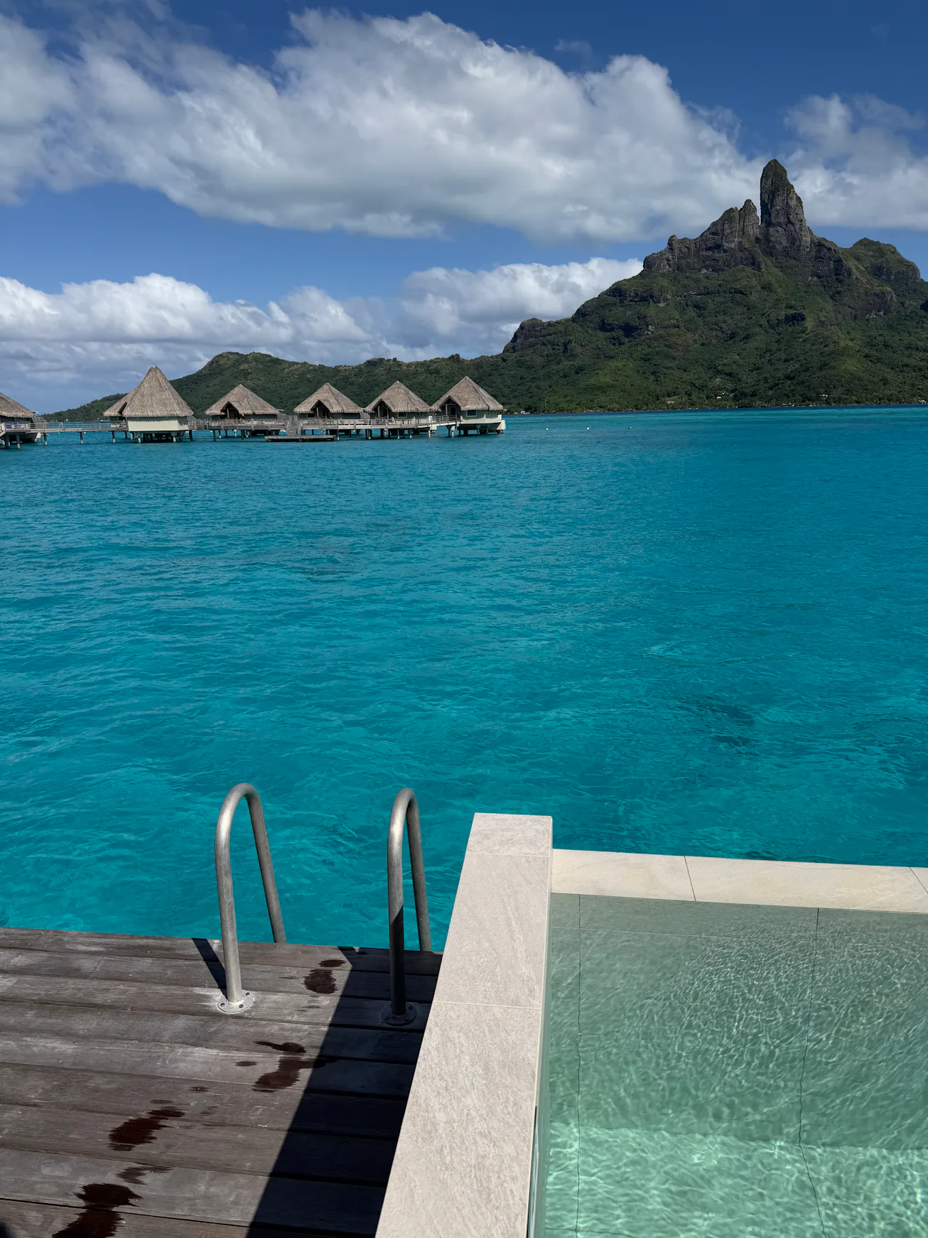 Turquoise lagoon with overwater bungalows on stilts, jagged volcanic island in background, wooden de...