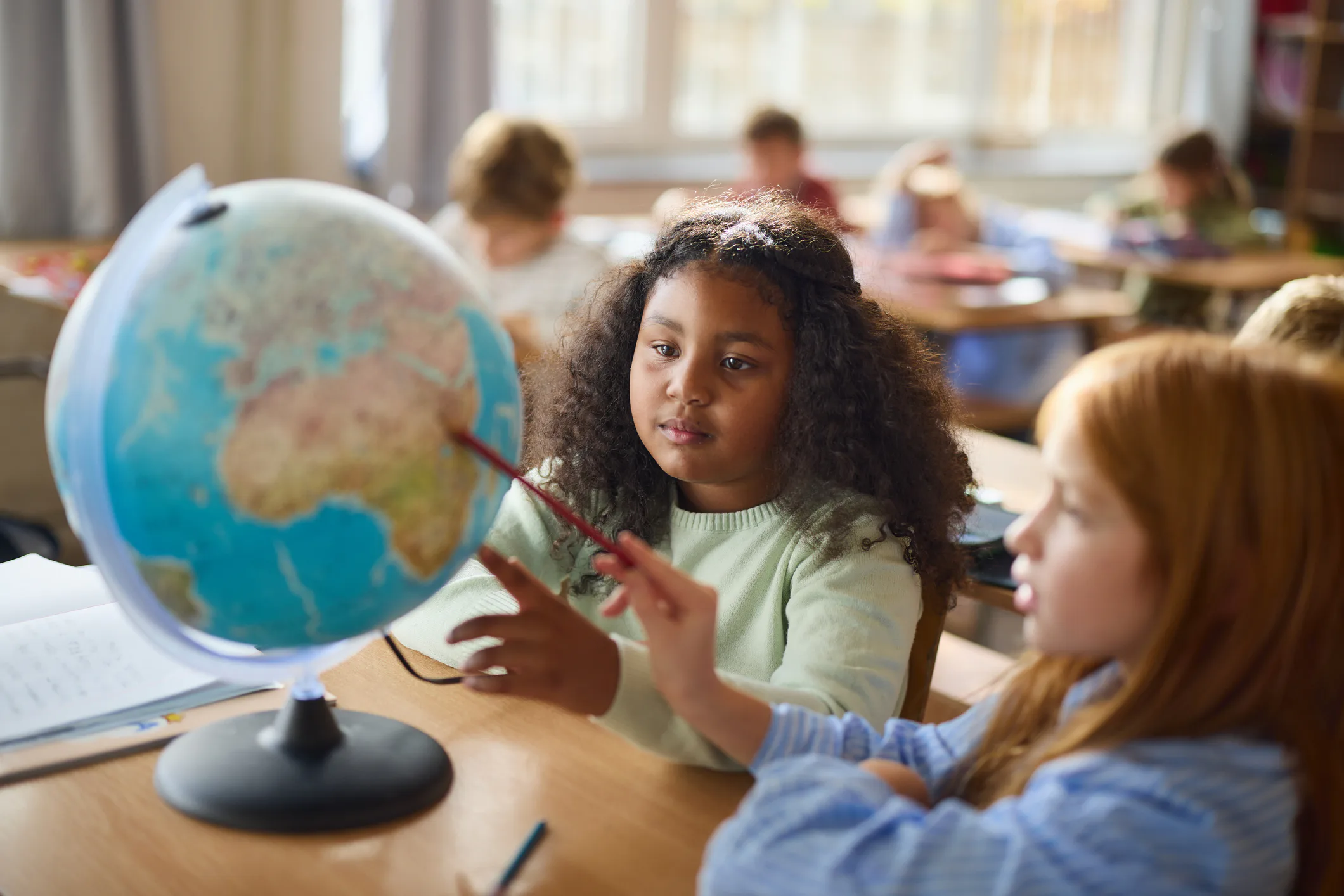 Two elementary students sit at a desk examining a tabletop globe; a girl with curly hair points with...