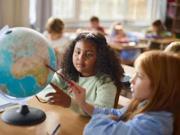 Two elementary students sit at a desk examining a tabletop globe; a girl with curly hair points with...