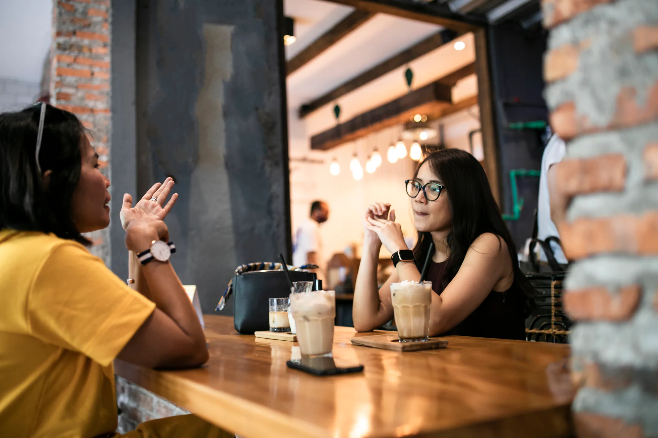 Two women sit across a wooden cafe table, each with iced coffee; one gestures while the other listen...