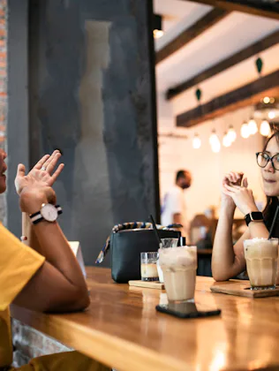 Two women sit across a wooden cafe table, each with iced coffee; one gestures while the other listen...