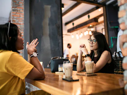 Two women sit across a wooden cafe table, each with iced coffee; one gestures while the other listen...