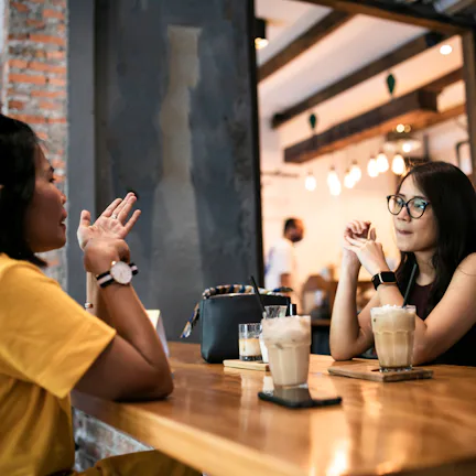 Two women sit across a wooden cafe table, each with iced coffee; one gestures while the other listen...