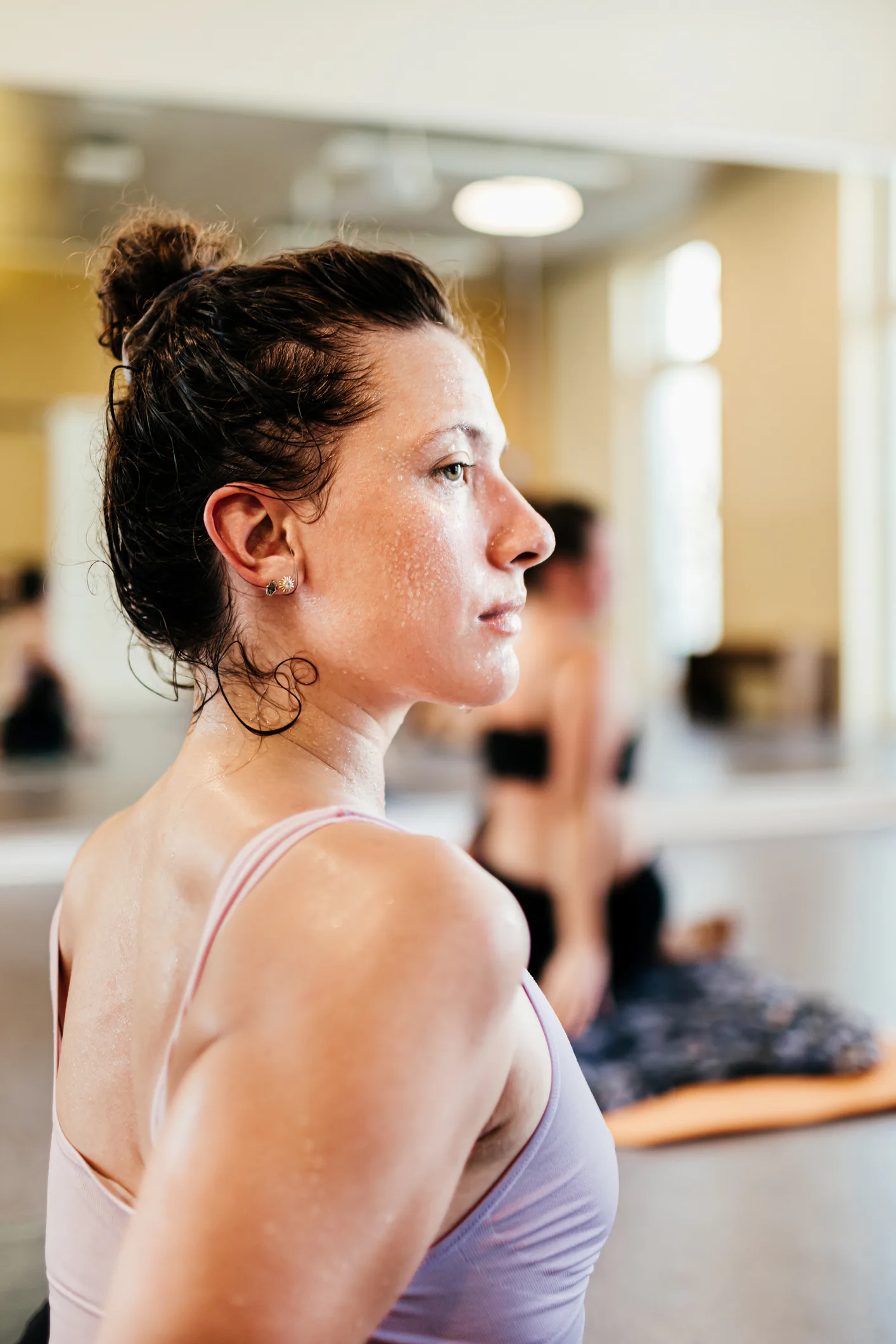 Side profile of a sweaty young woman in a sports bra with hair in a bun, looking focused in a bright...