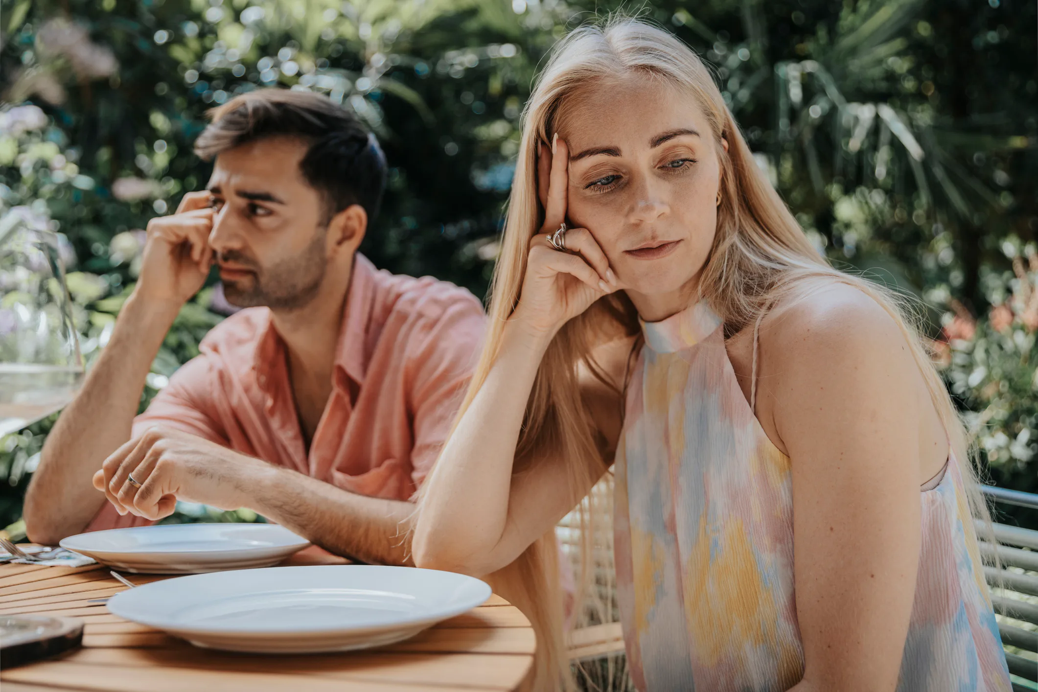 Two adults sitting at an outdoor table with empty plates, both looking away and appearing distant or...