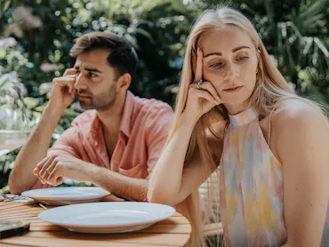 Two adults sitting at an outdoor table with empty plates, both looking away and appearing distant or...