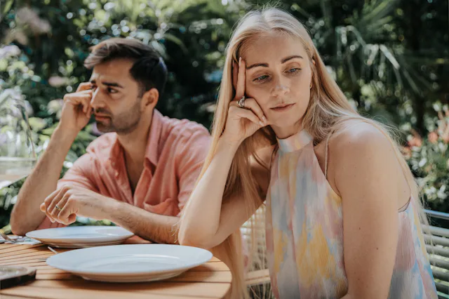 Two adults sitting at an outdoor table with empty plates, both looking away and appearing distant or...