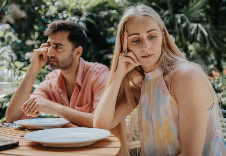Two adults sitting at an outdoor table with empty plates, both looking away and appearing distant or...