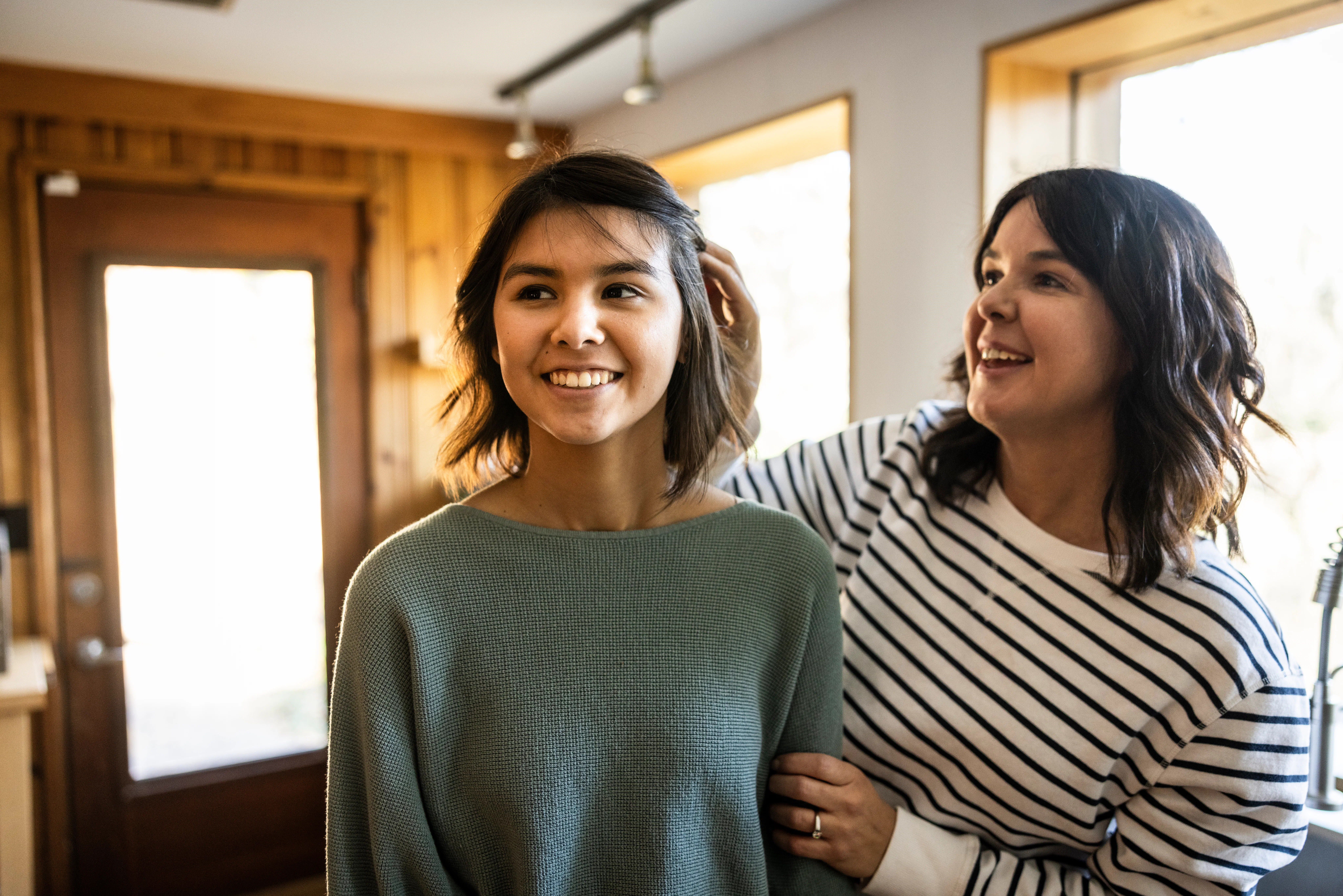 Two smiling women stand in a sunlit room near windows; one adjusts the other's hair while they chat,...