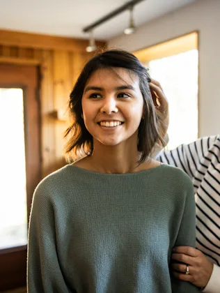 Two smiling women stand in a sunlit room near windows; one adjusts the other's hair while they chat,...
