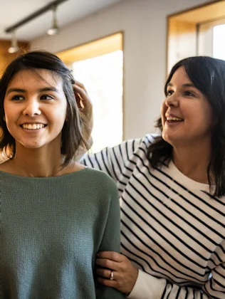 Two smiling women stand in a sunlit room near windows; one adjusts the other's hair while they chat,...