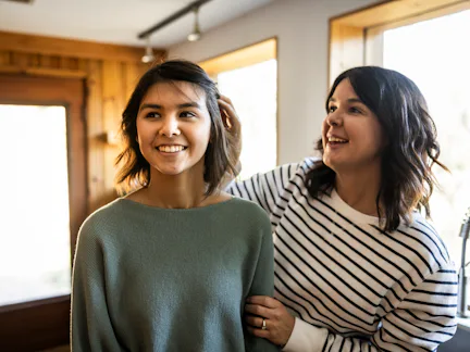 Two smiling women stand in a sunlit room near windows; one adjusts the other's hair while they chat,...